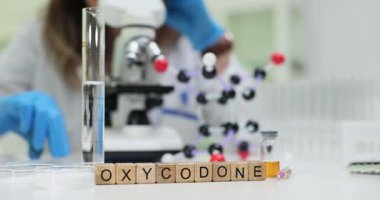 Tahta bloklar, cam tüp ve molekül modelinin yanında Oxycodone kelimesini heceler. Kadın araştırmacı, güçlü ağrı kesici riskini analiz ederek opioid örneğini kontrol ediyor.