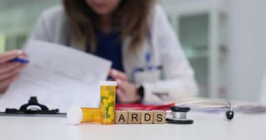 Doktor terapi önerilerini boş bir şekilde yazıyor. Minyatür ahşap bloklar ARDS kısaltması oluşturuyor. Uyuşturucu kapsüllerinin masasında duruyor.