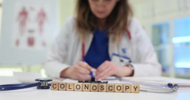 Kadın doktor kolonoskopi için tavsiye mektubunu tıbbi forma yazmış. Tahta bloklar düzenli inceleme masasına yerleştirilmiş. Kolonoskopi kelimesi.