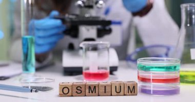 Kadın laboratuvar çalışanı, maddenin mikroskobik detaylarını inceliyor. Tahta küpler Osmium kelimesi oluşturur. Bu da kimyasal elementin soruşturma altında olduğunu gösterir.