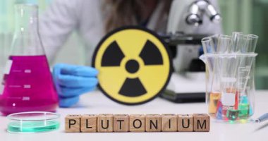 Laboratuvar çalışanı radyasyon tehlikesini işaret ediyor. Tahta küpler, bilimsel analizin tanımlayıcı konusu olan Plütonyum 'u oluşturur.