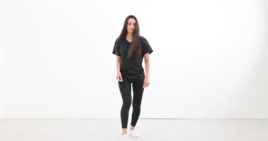 Tişört ve taytlı kadın geniş oturma odasında dans hareketleri yapıyor. Uzun saçlı bayan ağır çekim dans egzersizi yapıyor.