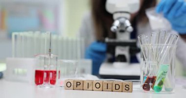 Tahta küpler, kan örnekleri ve cam eşyalar yanında afyon kelimesini heceler. Eldivenli laboratuar doktoru farmakoloji araştırması sırasında mikroskop altında nesneleri inceliyor.