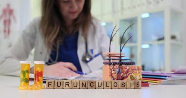 Tahta küplerde deri modeli ve ilaç şişelerinin yanında Furunculosis yazıyor. Mavi önlüklü kadın doktor hasta tedavisi hakkında notlar yazıyor.