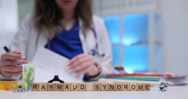 Kadın doktor, hasta için açık ve planlı bir terapi programı yazıyor. Masada minyatür tahta küplerle düzenlenmiş tıbbi terim Reynaud Sendromu.