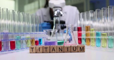 Kadın kimyager titanyum yapısını dikkatlice mikroskop altında inceliyor. Masadaki ahşap bloklar, laboratuvar masasında tüplerle Titanyum kelimesi oluşturur.