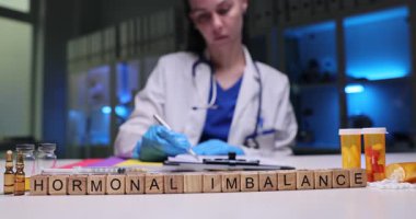 Beyaz önlüklü doktor hormonları dengelemek için talimatlar yazar. Masa başındaki tahta küplerin yanında hormon dengesizliği ifadesi var.