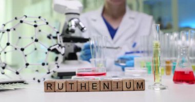 Kadın kimyager Petri kabında cam parçalarını dikkatlice inceler. Masanın üzerindeki minyatür tahta küpler araştırmanın odak noktasını gösteren kelime Rutenyum oluşturur