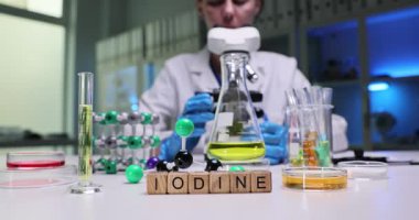 Kadın kimyager iyotu modern mikroskop altında dikkatlice inceliyor. Masanın üzerindeki minyatür ahşap küpler laboratuvar şişeleri ve moleküler şişelerle iyot oluşturur.