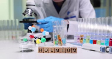 Laboratuvar çalışanı cam kaydırağa yerleştirilen çalışma örneklerini incelerken mikroskoba dikkatlice bakıyor. Tahta bloklar labaratuvar masasına yerleştirilmiş Holmium kelimesi.