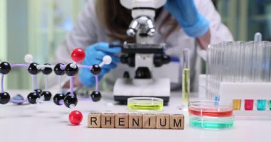 Masadaki küçük tahta küpler araştırma odağı olduğunu gösteren bir kelime olan Rhenium. Laboratuvar çalışanı, moleküler örnekleri incelemek için mikroskoba daha çok eğiliyor.
