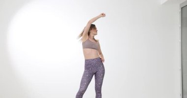 Spor sutyen ve taytlı genç bir kadın parlak stüdyoda enerjik bir şekilde dans ediyor. Kadın hareketli adımlar atıyor. Kalp ritmi eğitimi alırken kolunu kaldırıyor.