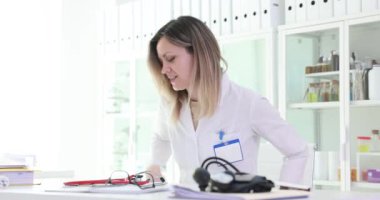 Kadın doktor masasına oturmuş masadan steteskop ve gözlük kaldırıyor. Kadın, vücudu yoğun hastane vardiyasına hazırlarken burun çerçevelerini ayarlıyor.