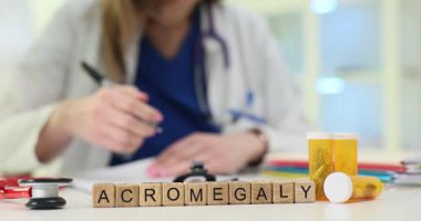 Tıbbi masadaki tahta bloklar ilaç kutularının yanında Acromegaly kelimesini heceliyor. Kadın doktor hasta grafiği üzerinde büyüme bozukluğu üzerine notlar yazıyor.