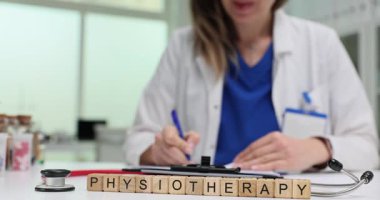 Tıbbi masadaki tahta bloklar stetoskop yakınlarındaki fizik tedavi kelimelerini heceliyor. Kadın doktor hasta için fizik tedavi seansı raporu yazıyor.