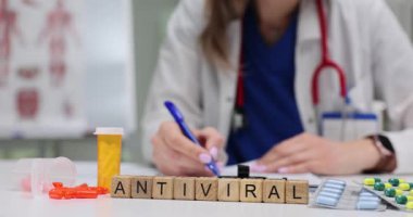 Tahta bloklar klinik masasında antiviral kelimesini heceliyor. Kadın doktor randevuda virüs tedavisi için ilaç hazırlarken reçete yazar