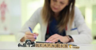 Masadaki tahta bloklar otoskop cihazının yanında sağırlık kelimesini heceliyor. Kadın doktor, hastanın işitme testi notlarını okurken aynı zamanda işitme testi notlarını da yazıyor.