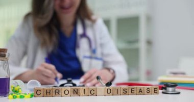 Tahta bloklar klinik masasında Kronik Hastalık kelimelerini heceliyor. Kadın doktor randevuda hasta için uzun vadeli tedavi notları yazıyor.