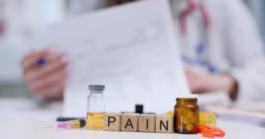 Masadaki tahta bloklar şişelerin ve şırıngaların yanında acı sözcüğü heceliyor. Kadın doktor sürekli ağrı kontrol planı hakkında detaylı not yazıyor.