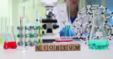 Kadın kimyager Niobium 'u inceliyor ve not defterine mikroskop ile not alıyor. Tahta küpler test tüplerinin yanındaki masada çözeltilerle Niobiyum kelimesi oluşturur.