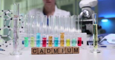 Kadın kimyager kimyasalları karıştırır ve gözlemleri titizlikle kaydeder. Tahta küpler test tüplerinin yanında kelime kadmiyumu oluşturur ve raflarda renkli sıvılar bulunur.