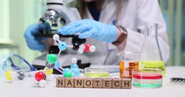 Kadın laboratuvar teknisyeni mikroskop kullanarak hassas örnek yapılarını inceliyor. Tahta küpler, laboratuvar masasına güzelce yerleştirilmiş. Camda Nanoteknoloji sözcüğü.
