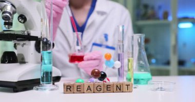 Laboratuvar çalışanı şeffaf cam deney şişesi tutuyor ve analiz için canlı kırmızı sıvı çizmek için pipetini kullanıyor. Tahta küpler kelime ayıracı oluşturmak için düzenlenmiş