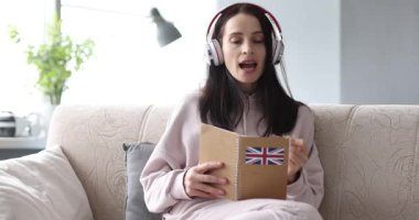 Kulaklık takan kadın kanepeye oturup kitap okuyor ve kapağında İngiliz bayrağı var. Öğrenci sayfadaki yeni kelimeleri tekrarlayarak İngilizce telaffuz pratiği yapar