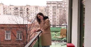 Bej ceketli neşeli kadın balkonda duruyor ve telefonda konuşuyor. Şık bir kadın güzel şehir manzarasına hayran. Bayan telefonda sıcak bir şekilde konuşuyor.