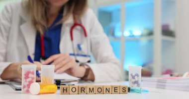 Kadın doktor test sonuçlarını titizlikle inceler ve notlar alır. Tahta küpler hasta için ilaç kutularının yanındaki masada kelime hormonu oluşturur.