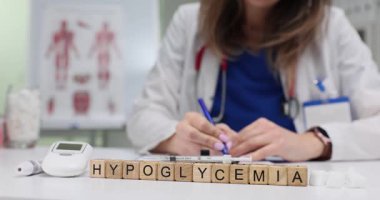 Masadaki tahta bloklar kelime hipoglisemiyi heceliyor. Glukometre ve şeker küplerinin yanında. Mavi önlüklü doktor kan şekerini ölçen notlar yazıyor.