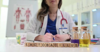 Ahşap bloklar, cam şişelerin yanında renkli sıvıyla Lactobacillus kelimesini heceler. Kadın doktor probiyotik bakteri ve mide dengesi hakkında notlar yazıyor.