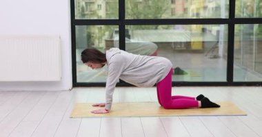 Kapüşonlu genç kadın kalça ve karın kasları için yer egzersizi yapıyor. Sporcu bacağını vücudunun gerisine kaldırıyor. Elleri ve dizlerini odada paspasın üzerinde tutuyor.