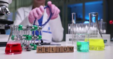 Masa üzerindeki tahta bloklar renkli reaksiyonların yanında Helyum kelimesi. Kimya bilimcisi deney tüpünün yanında gözlük ve laboratuvarda ağır çekimde mataralar tutuyor.