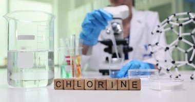 Masadaki tahta küpler temiz beher ve rafın yanında klor kelimesini heceliyor. Laboratuvar çalışanı mikroskobun yanında petri kabına sıvı pipetler.