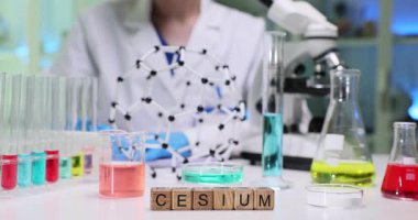 Tahta bloklar laboratuvar masasında sezyum kelimesini heceler. Palto giyen bilim adamı cam ve mikroskop kullanarak renkli test tüpleri ve moleküler modeller üzerinde çalışıyor.