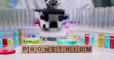 Tahta bloklar mikroskop ve test tüplerinin önünde Prometyum kelimesini heceler. Beyaz önlüklü bilim adamı renkli sıvıları incelerken örnekler hazırlıyor.