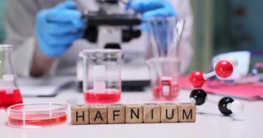 Tahta bloklar Hafniyum kelimesini parlak laboratuvar masasında heceler. Eldiven bilimcileri mikroskobu ayarlıyor. Kırmızı sıvıyla beherlerde örnekler hazırlıyorlar.