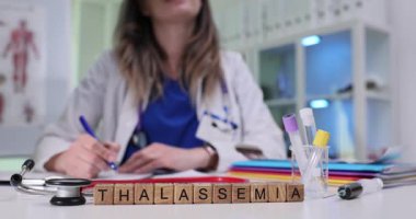 Tahta bloklar Thalassemia kelimesini test tüpleri ve steteskop yakınında laboratuvar masasında hecelerler. Kadın doktor anemi yönetimini inceleyen hematoloji notları yazıyor.