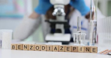 Masadaki tahta bloklar cam eşyanın yanında Benzodiazepine diye okunuyor. Kadın kimyagerler mikroskop kullanarak sakinleştirici ilaç örneklerini değerlendiriyor.