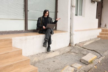 Genç bir kadın bir çıkıntıda oturuyor, dizüstü bilgisayarıyla meşgul açık gökyüzünün altında çalışıyor. Şehir hayatı vızıldarken güneş ışığı odaklanmış ifadesini vurgular..