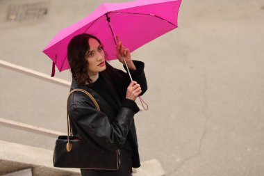 Bir kadın elinde parlak pembe bir şemsiyeyle beton merdivenlerde duruyor. Fırtınalı bir şehirde etrafındaki yağmur damlaları gibi derin düşünceler içinde yukarı bakıyor. Atmosfer sakin ama yansıtıcı..