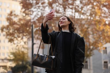 Bir kadın, şehir parkında dururken akıllı telefonuyla selfie çekiyor. Altın yapraklar nazikçe etrafını sarıyor ve ılık bir sonbahar havası yaratıyor..