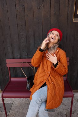 Parlak turuncu ceketli ve kırmızı şapkalı bir kadın telefonda konuşurken gülüyor. Neşe dolu bir sahneyi güzelleştiren ahşap bir arka planla bir bankta eğleniyor..