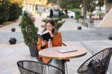 Açık havada hareketli bir kafede, kitaplarına dalmış bir kadın, insanlar yürürken ve ağaçlar altın rengine dönerken serin sonbahar havasının tadını çıkarıyor. Yakınlarda bir şişe su duruyor, dinginliğini arttırıyor..