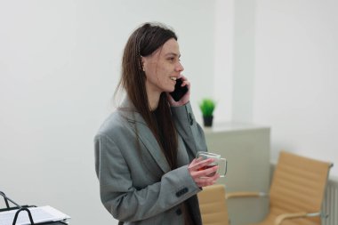 Genç bir profesyonel telefonda konuşurken gülümsüyor, parlak, minimalist bir ofiste bir bardak tutuyor. Doğal ışık boşlukta akıyor, pozitif titreşimi arttırıyor..