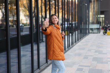Bir kadın modern bir binanın dışında selfie çekerken ışıl ışıl gülümser. Sıradan mavi kotların üzerine göz alıcı turuncu bir ceket giyer..