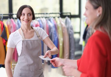 Renkli bir giyim mağazasında, neşeli bir tezgahtar memnun bir müşteriye makbuzu verirken gülümsüyor. Parlak giysiler arka planda asılı, canlı bir atmosfer yaratıyor..