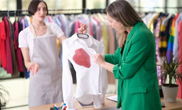 Kalabalık bir giyim mağazasında, müşteri beyaz bir gömlekte kırmızı bir leke bulur. Tezgahtar endişeli bir şekilde bakıyor. Parlak giysiler çevrelerini sarar, canlı bir atmosfer yaratır..