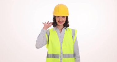 Gülümseyen kadın mühendis güvenlik yeleği ve sarı kask giyiyor. İnşaat uzmanı tamamlanmış proje kalitesini onaylayan el işaretini gösterdi